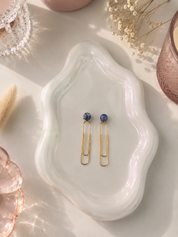Gold earrings with blue stones on a white ceramic dish with dried flowers and a candle.