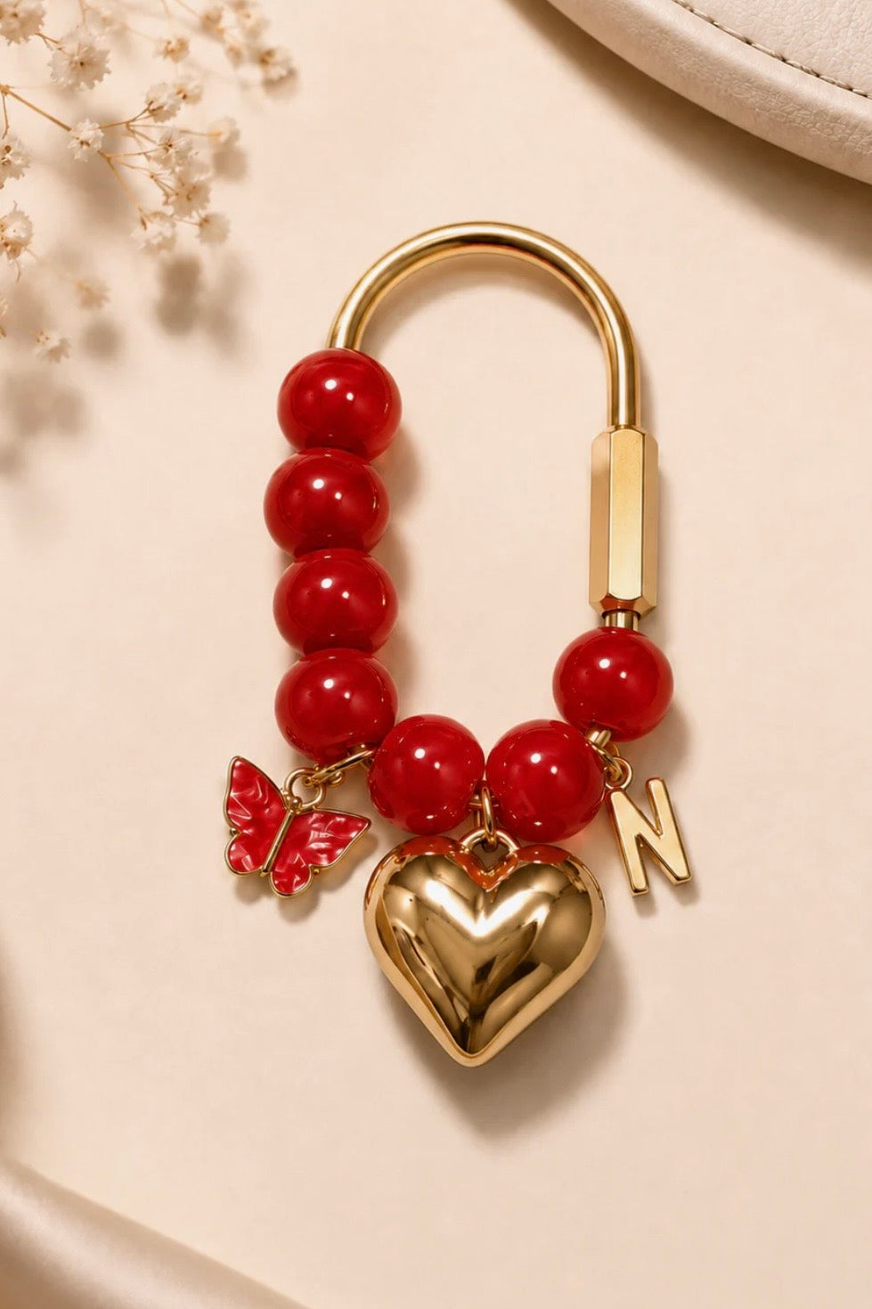 A gold-tone and red beaded charm keychain featuring a heart-shaped locket, a butterfly charm, and a letter 'N' charm, displayed on a beige background with dried flowers.