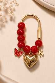 A gold-tone and red beaded charm keychain featuring a heart-shaped locket, a butterfly charm, and a letter 'N' charm, displayed on a beige background with dried flowers.