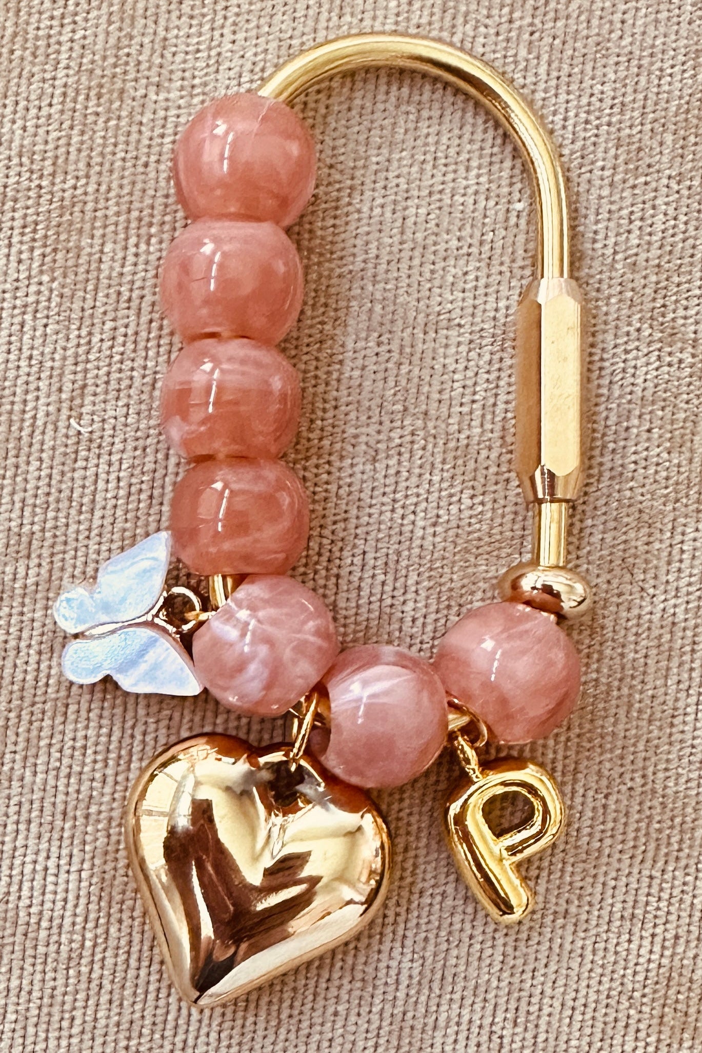 A pink stone bead keychain with gold-tone metal hardware, featuring a polished gold heart charm, a small gold letter 'P' charm, a white butterfly charm, and a clasp on a beige textured fabric background.