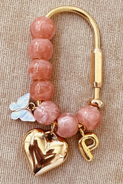 A pink stone bead keychain with gold-tone metal hardware, featuring a polished gold heart charm, a small gold letter 'P' charm, a white butterfly charm, and a clasp on a beige textured fabric background.