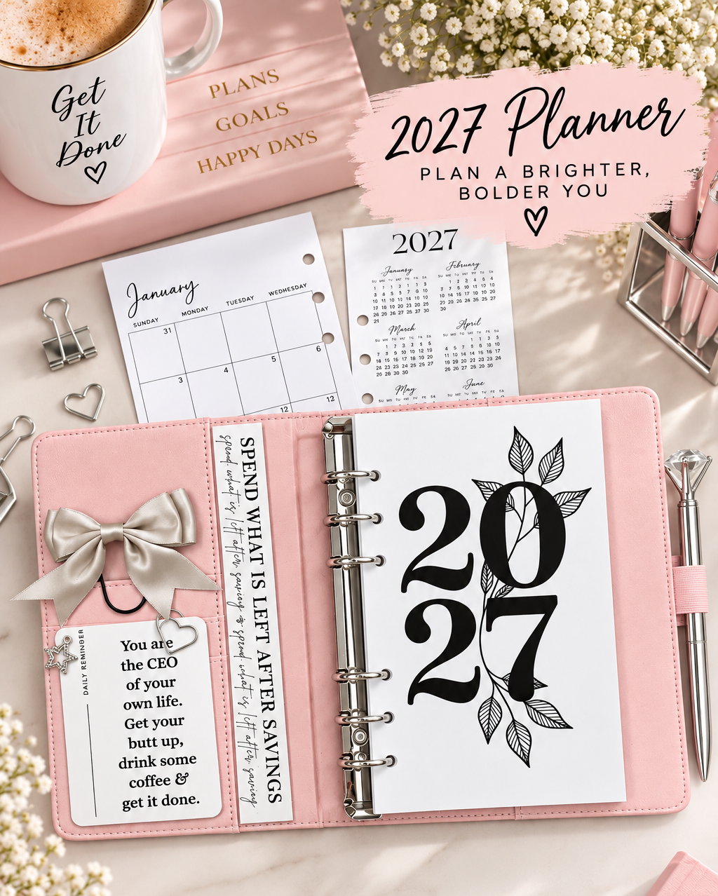 A pink ring-bound 2027 planner shown open on a light marble surface with stationery props, including a mug, clips, pen, and additional paper inserts. The main product is the planner cover and binder, with large black '2027' numerals and leaf graphics on the page.