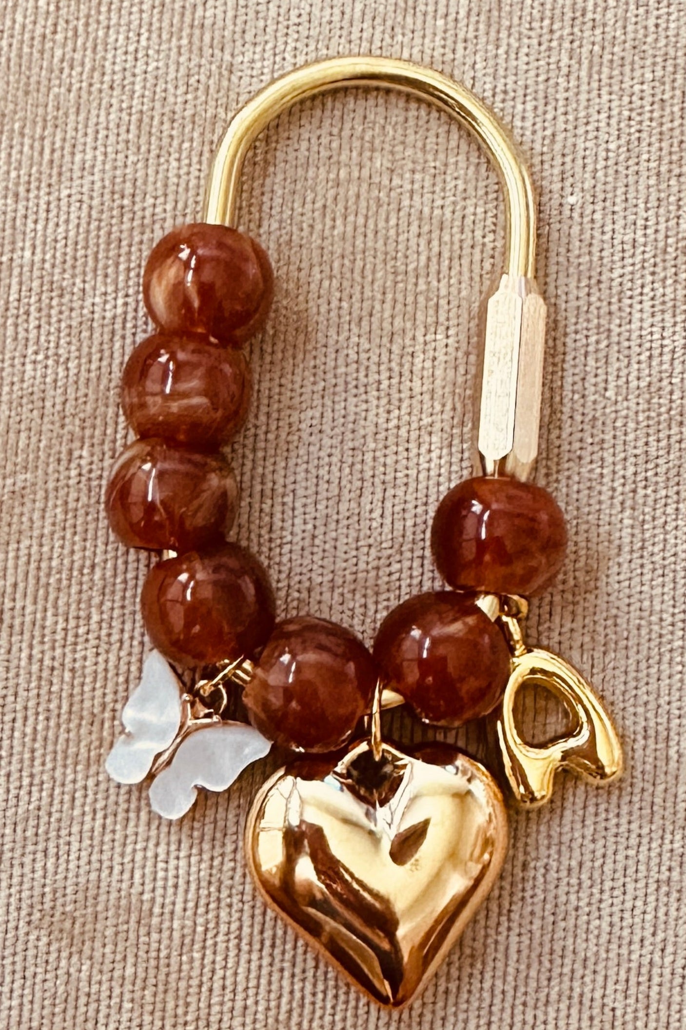 A beaded charm bracelet with brown translucent beads, a gold-tone metal clasp, a polished gold heart charm, and a small white butterfly charm, displayed on a beige textured fabric surface.