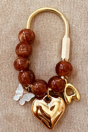 A beaded charm bracelet with brown translucent beads, a gold-tone metal clasp, a polished gold heart charm, and a small white butterfly charm, displayed on a beige textured fabric surface.