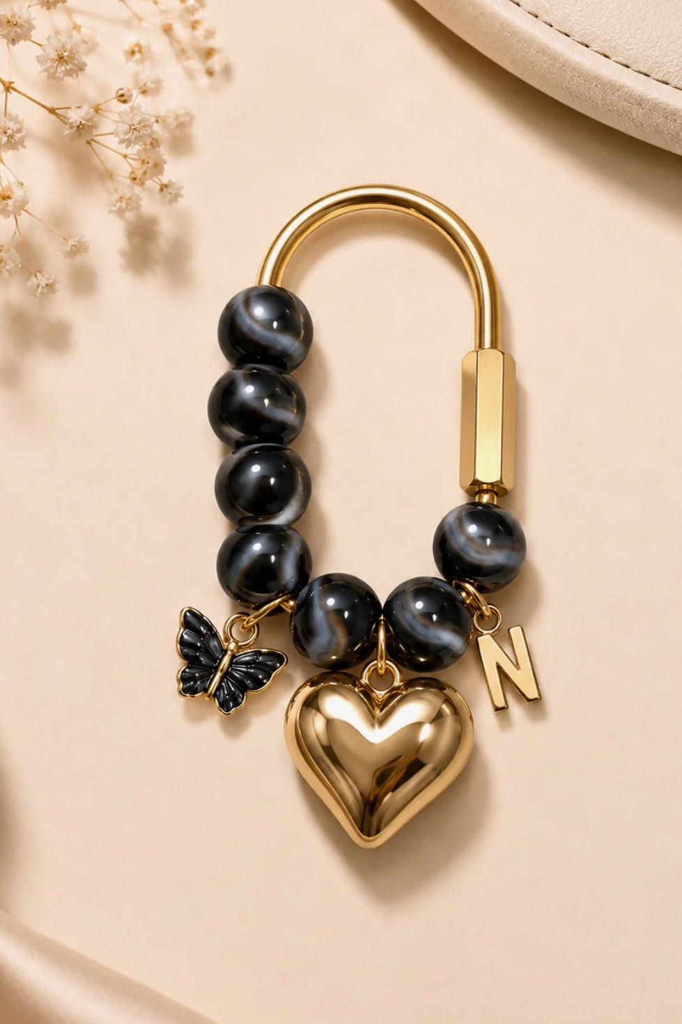 A gold-tone keychain or bracelet charm featuring dark gray tiger-eye beads, a polished gold heart, a butterfly charm, and a letter 'N' charm, displayed on a beige satin background.
