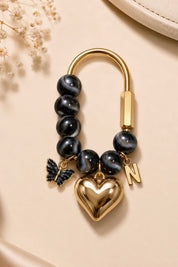 A gold-tone keychain or bracelet charm featuring dark gray tiger-eye beads, a polished gold heart, a butterfly charm, and a letter 'N' charm, displayed on a beige satin background.