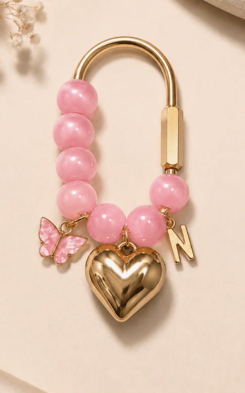 A gold-tone keychain or bag charm featuring a series of pink beads, a polished gold heart pendant, a pink butterfly charm, and a gold initial 'N' charm, displayed on a light beige background.