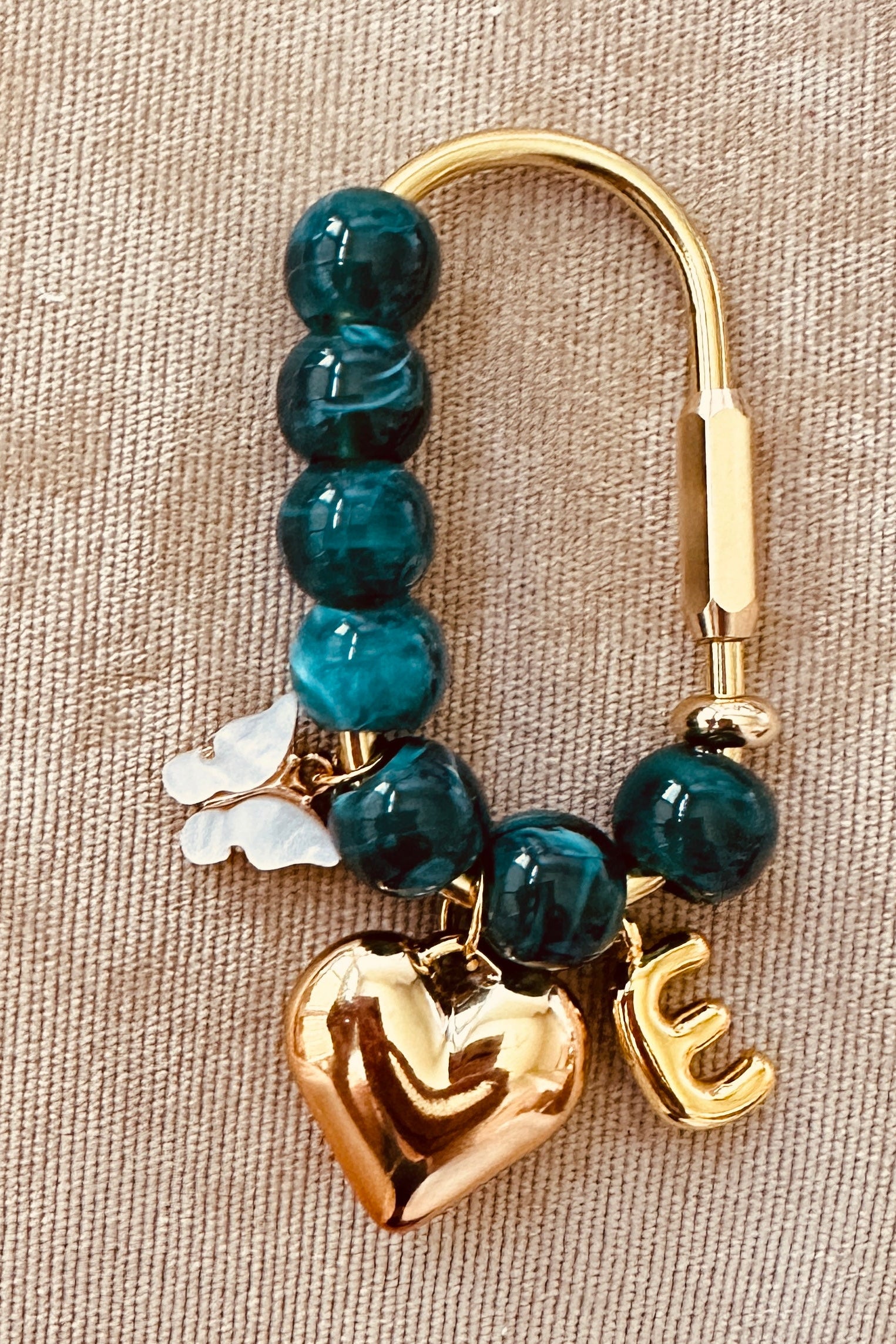 A gold-tone clasp keychain or bag charm featuring round teal beads, a polished gold heart charm, a gold letter 'E' charm, and a small white flower charm, displayed on a textured beige fabric background.