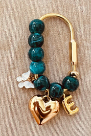 A gold-tone clasp keychain or bag charm featuring round teal beads, a polished gold heart charm, a gold letter 'E' charm, and a small white flower charm, displayed on a textured beige fabric background.