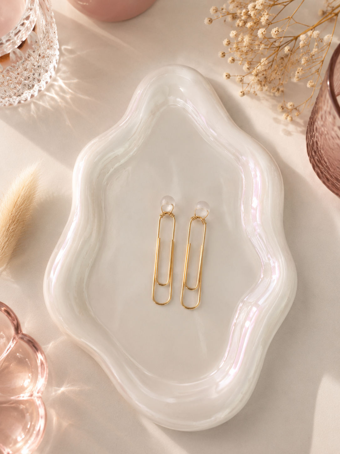 Gold earrings on a white wavy dish with dried flowers and a brush in the background
