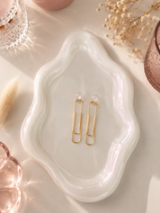 Gold earrings on a white wavy dish with dried flowers and a brush in the background