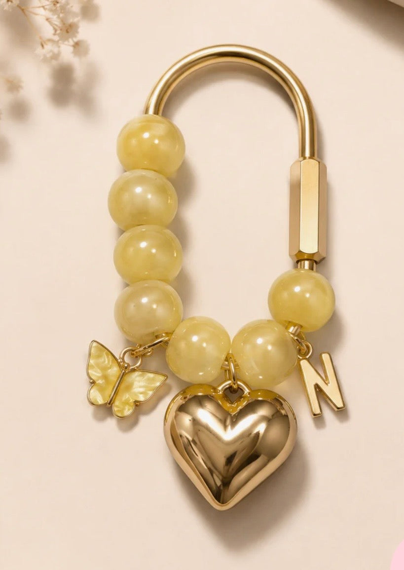 A gold-toned keychain or bag charm featuring round pale yellow beads, a shiny gold heart pendant, a butterfly charm, and a letter 'N' charm, displayed on a light beige background.