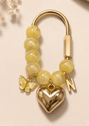 A gold-toned keychain or bag charm featuring round pale yellow beads, a shiny gold heart pendant, a butterfly charm, and a letter 'N' charm, displayed on a light beige background.