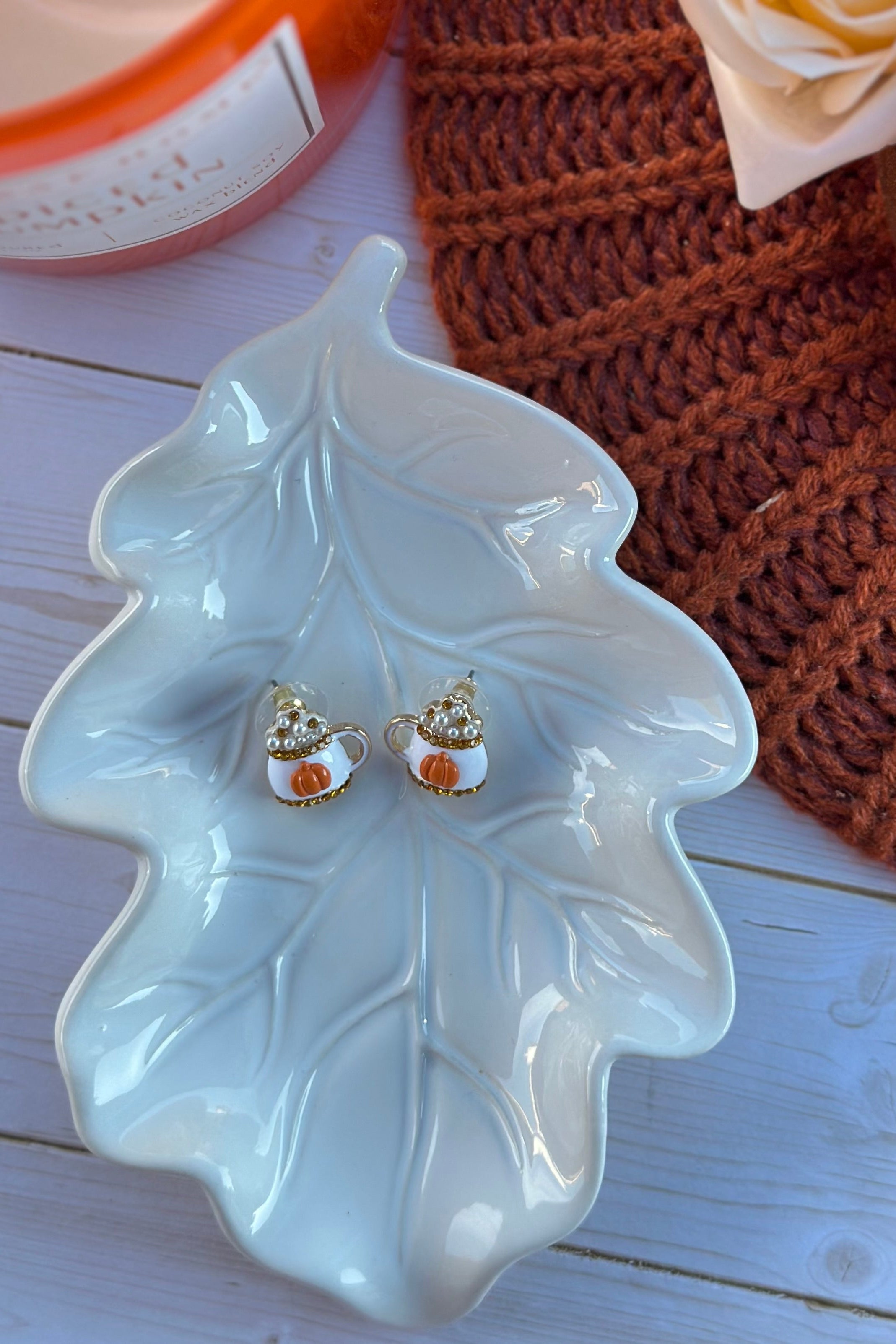 Pair of earrings on a leaf-shaped ceramic dish with a brown knitted item in the background.