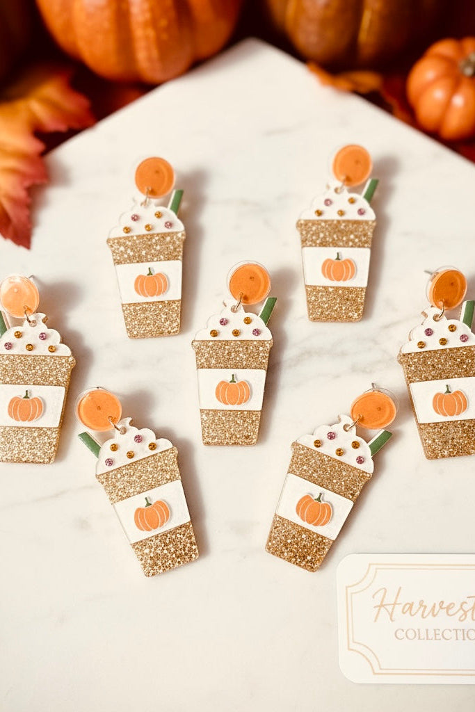Pumpkin spice latte earrings on a white surface with pumpkins and leaves in the background.