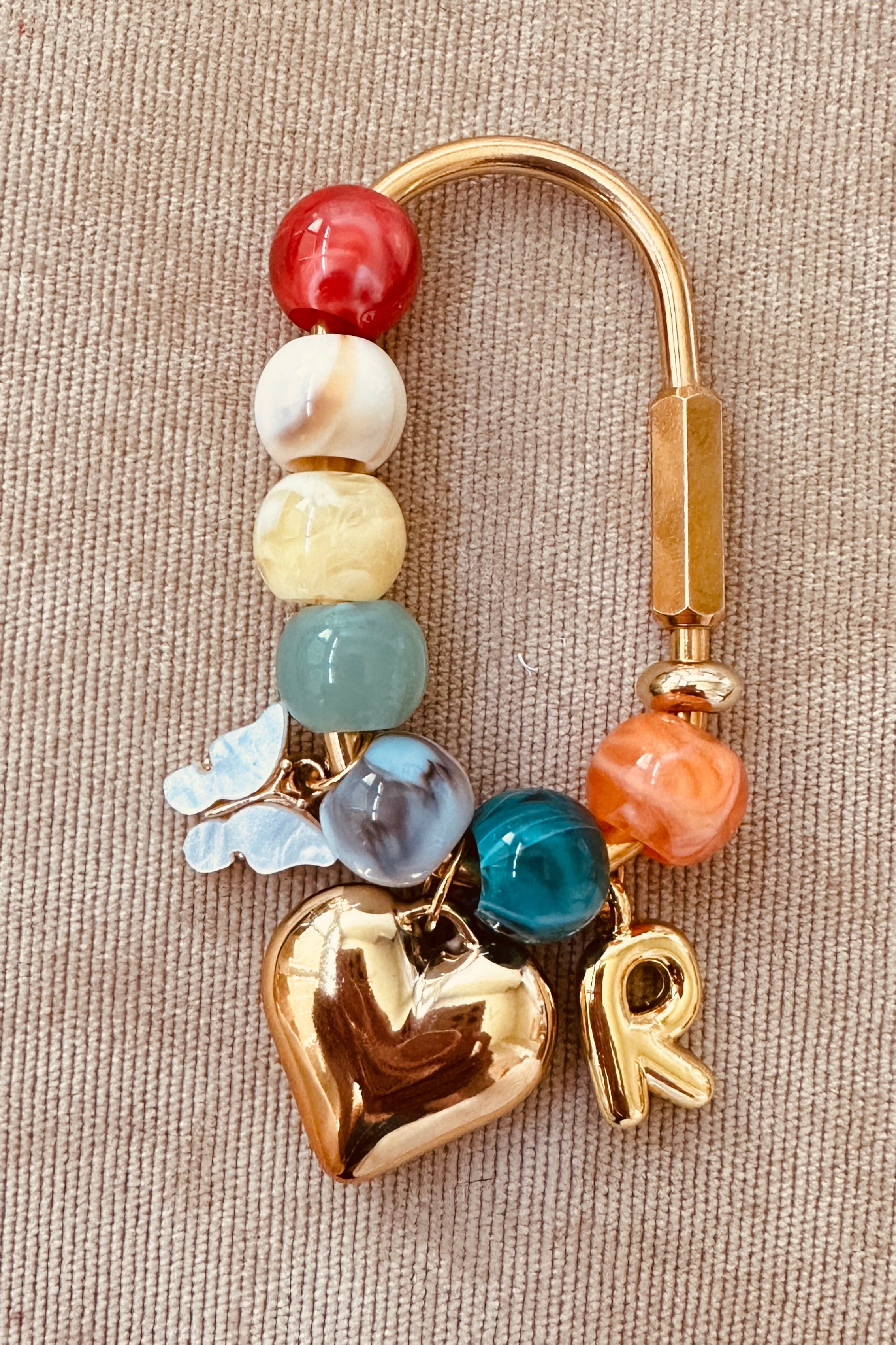 A gold-toned beaded charm accessory with a heart-shaped charm, a letter 'R' charm, and colorful marbled beads, displayed on a textured beige fabric background.