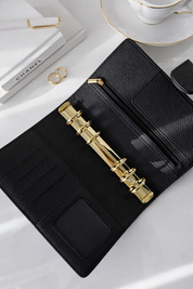 Black leather wallet with gold accents on a white surface, with Chanel books and a teacup in the background.