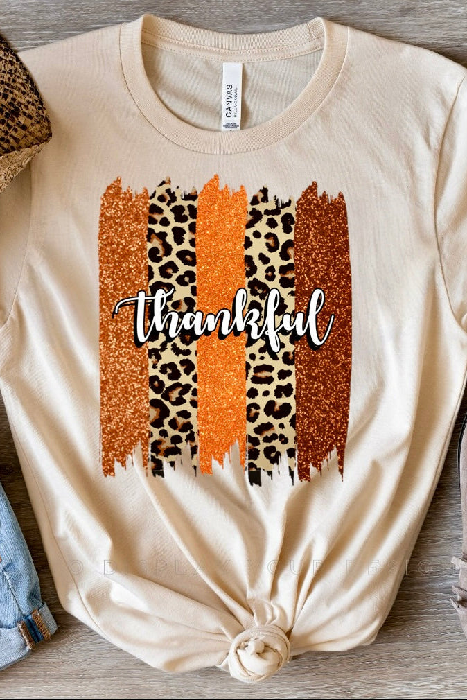A beige t-shirt with leopard print and the word 'THANKFUL' printed on it, displayed flat with a hat, sunglasses, and shoes arranged around it.