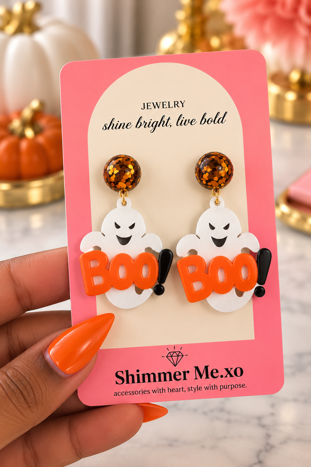 A hand with orange nail polish holds a pink display card featuring a pair of Halloween-themed dangling earrings. The earrings have glittery amber-brown round studs and white ghost charms with black eyes holding bright orange 'BOO' lettering.