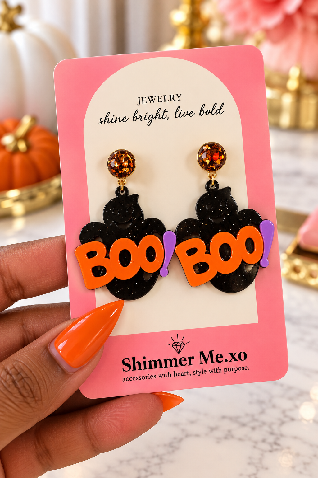 A hand with orange nails holds a pink display card featuring a pair of Halloween earrings. The earrings have black glittery ghost shapes with orange 'BOO' lettering and purple exclamation marks, with gold-toned studs topped by round orange glitter beads.