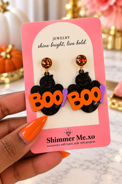 A hand with orange nails holds a pink display card featuring a pair of Halloween earrings. The earrings have black glittery ghost shapes with orange 'BOO' lettering and purple exclamation marks, with gold-toned studs topped by round orange glitter beads.