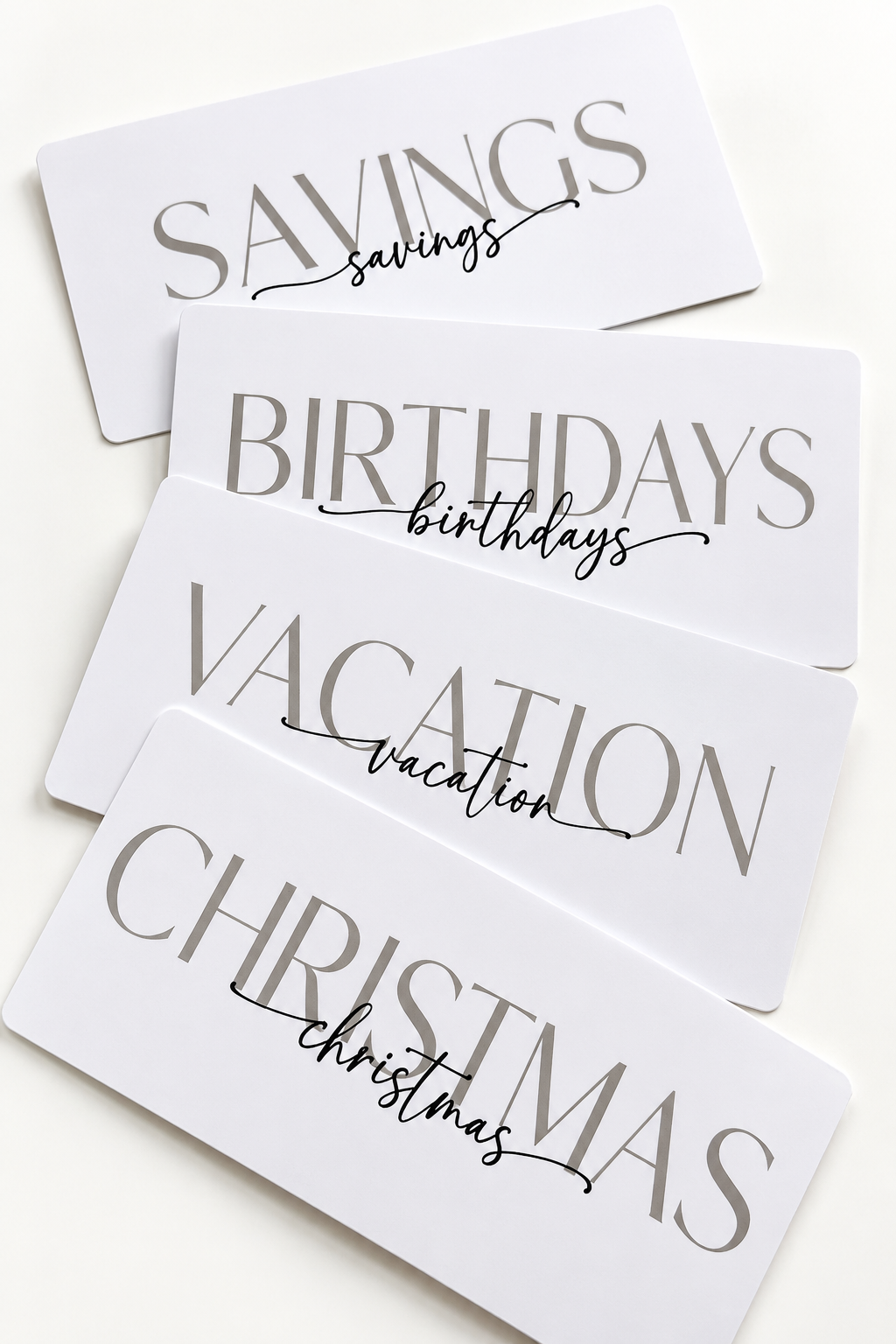 Five white rectangular gift cards with elegant gray typography, stacked diagonally on a light cream background. The cards read 'SAVINGS', 'BIRTHDAYS', 'VACATION', and 'CHRISTMAS', with 'savings', 'birthdays', 'vacation', and 'christmas' written in a delicate black script underneath.