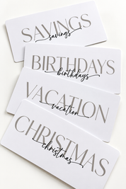 Five white rectangular gift cards with elegant gray typography, stacked diagonally on a light cream background. The cards read 'SAVINGS', 'BIRTHDAYS', 'VACATION', and 'CHRISTMAS', with 'savings', 'birthdays', 'vacation', and 'christmas' written in a delicate black script underneath.