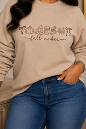 A woman wearing a beige crewneck sweatshirt with a brown graphic that reads 'fall vibe' with autumn-themed icons, paired with blue jeans, posing in a cozy indoor setting.