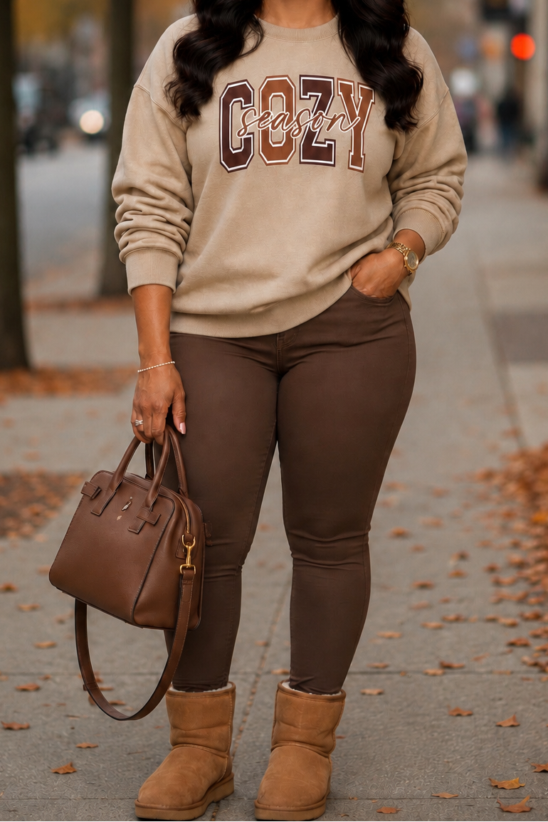 A woman stands on an autumn city sidewalk wearing a beige sweatshirt with 'COZY' in brown and burgundy varsity letters, brown leggings, tan boots, and carrying a brown handbag.