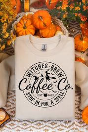 White sweatshirt with 'Witches Brew Coffee Co' print on a festive background with pumpkins and flowers.